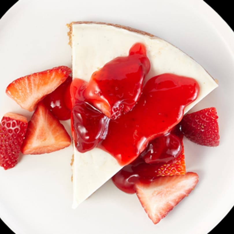 A slice of cheesecake with strawberry topping and fresh strawberry pieces on a white plate.